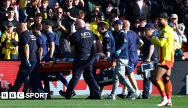 Footballer Conor Coady being carried off a football pitch on a stretcher by a team of male medics wearing dark colours while a player in a yellow shirt with red trim, red shorts, white socks and white boots is visible in the foreground and fans applauding are visible in the background