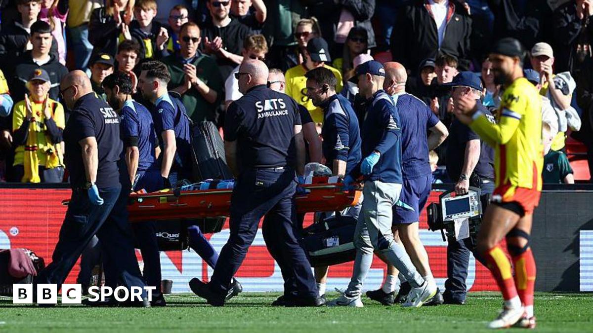 Footballer Conor Coady being carried off a football pitch on a stretcher by a team of male medics wearing dark colours while a player in a yellow shirt with red trim, red shorts, white socks and white boots is visible in the foreground and fans applauding are visible in the background
