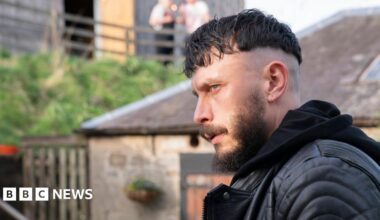 Richard Gadd stares intensely. He is wearing a black leather jacket and hoodie and is outside. Stone and slate buildings can be seen behind him. There are two people far behind on a terrace.