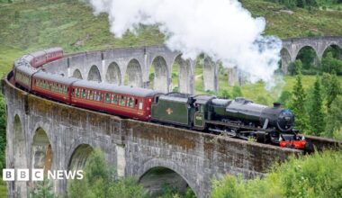 Steam rises from the train's black locomotive as it crosses the Glenfinnan Viaduct. The locomotive is hauling a line of carriages.