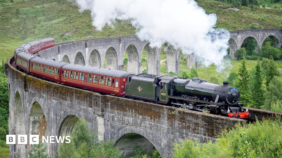 Steam rises from the train's black locomotive as it crosses the Glenfinnan Viaduct. The locomotive is hauling a line of carriages.