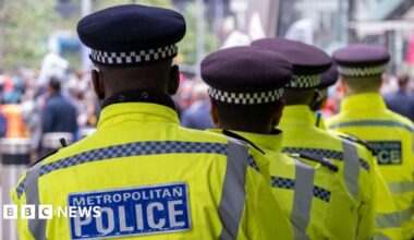 Five officers stand in a row in their yellow and blue uniforms. Each wears a black hat and stand looking out to a crowd of people.