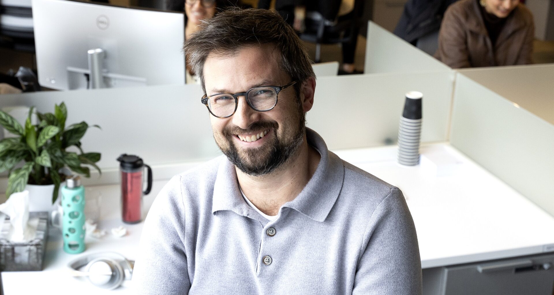 Jonathan Guillemette smiles while standing in an office in front of other employees doing work.