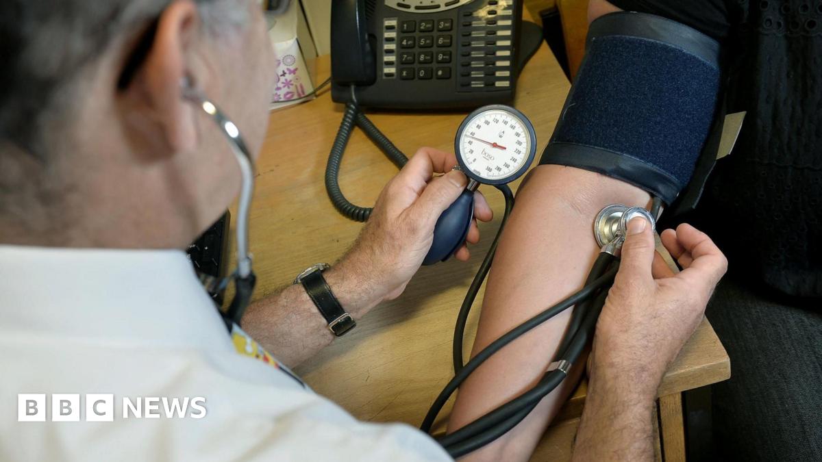 A doctor conducting a blood pressure test on a patient. He is holding a medical instrument against an arm whilst monitoring a gauge. The patient has a dark blue wrap around their arm.