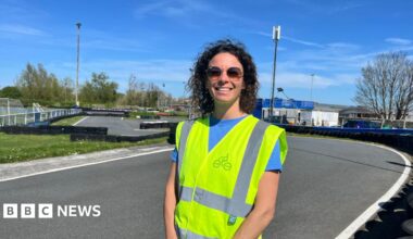 A lady in a high viz yellow jacket stands on a tarmac road.  She's wearing sunglasses with a dark blue sky in the background.