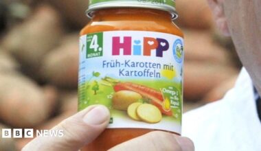 A man holds a jar of HiPP carrot and potato baby food, with potatoes stacked in the background.