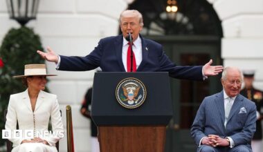 Donald Trump is standing at a lectern featuring the official coat of arms of the US presidency. He has both arms raised out to the sides. King Charles is sitting at one side and Trump's wife Melania is on the other. The King is wearing a light blue pin-striped suit with a light shirt and a light blue tie with a light blue pocket square. He is looking to the side and laughing. Trump is wearing a dark blue suit jacket, white shirt and red tie. He is mid speech. Melania is wearing a white fitted suit and a straw hat. They are outside the White House