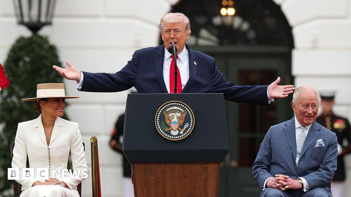 Donald Trump is standing at a lectern featuring the official coat of arms of the US presidency. He has both arms raised out to the sides. King Charles is sitting at one side and Trump's wife Melania is on the other. The King is wearing a light blue pin-striped suit with a light shirt and a light blue tie with a light blue pocket square. He is looking to the side and laughing. Trump is wearing a dark blue suit jacket, white shirt and red tie. He is mid speech. Melania is wearing a white fitted suit and a straw hat. They are outside the White House