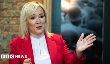 Two girls smiling at the camera as they pose for a picture in a youth club. Maisie on the left has brown hair pulled back in a pony tail and is wearing a grey hoodie with Harvard University branding in large red type. Iga on the right wears a black hoodie bunched up on her arms. She has shoulder-length blond hair and has rimless glasses.