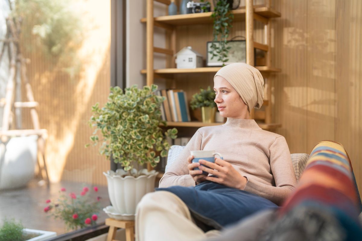 A young woman with cancer sits quietly by the window in a bright, warm room, sipping on a warm drink. The space is cozy with plants, light-colored wooden furniture, and plenty of natural light.
