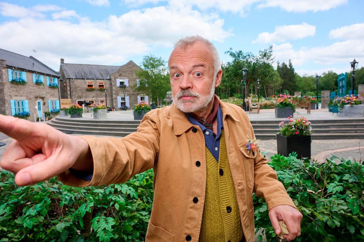 An older individual, dressed in a light brown jacket, stands in an outdoor setting and points towards an unspecified direction, with various buildings and greenery visible in the background.