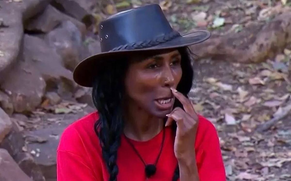 An individual dressed in a red shirt and a wide-brimmed black hat, seated outdoors with one hand to their face, appears to be engaged in a contemplative or reflective pose amidst a rocky environment.