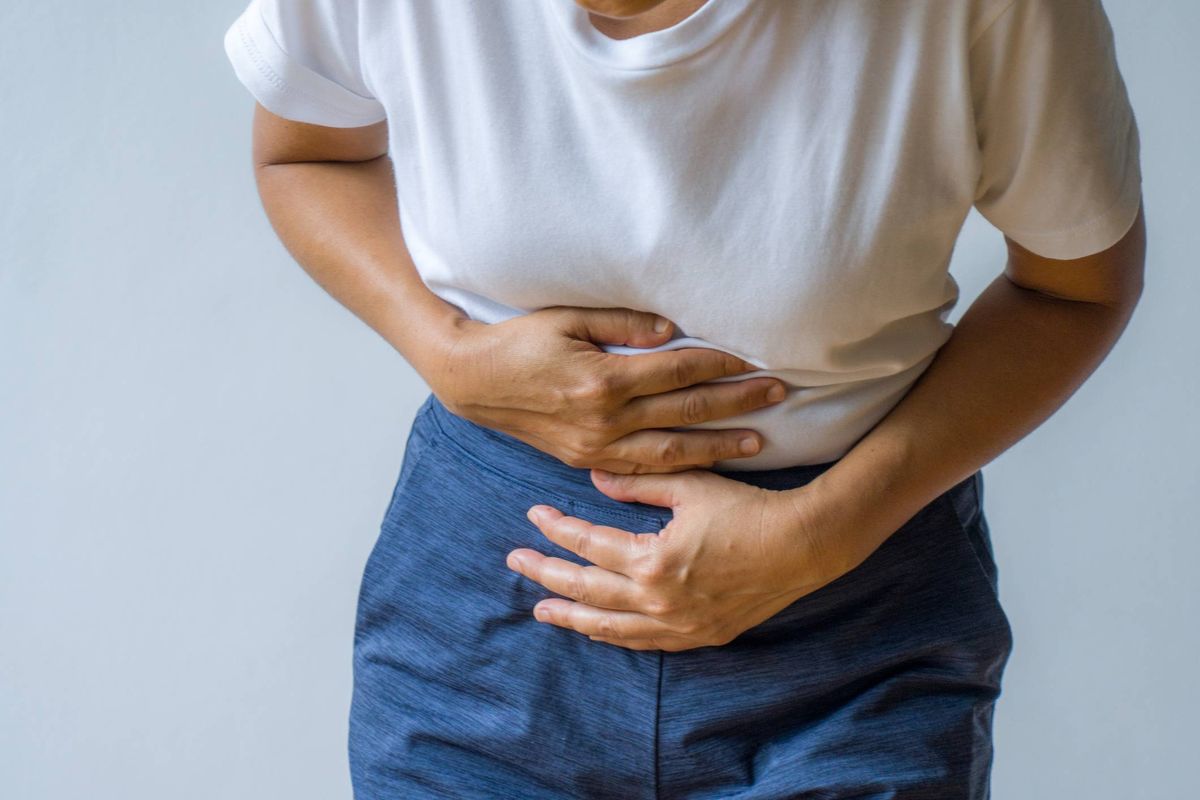 An individual dressed in a white shirt and blue jeans appears to be experiencing discomfort, with both hands placed on their abdomen.
