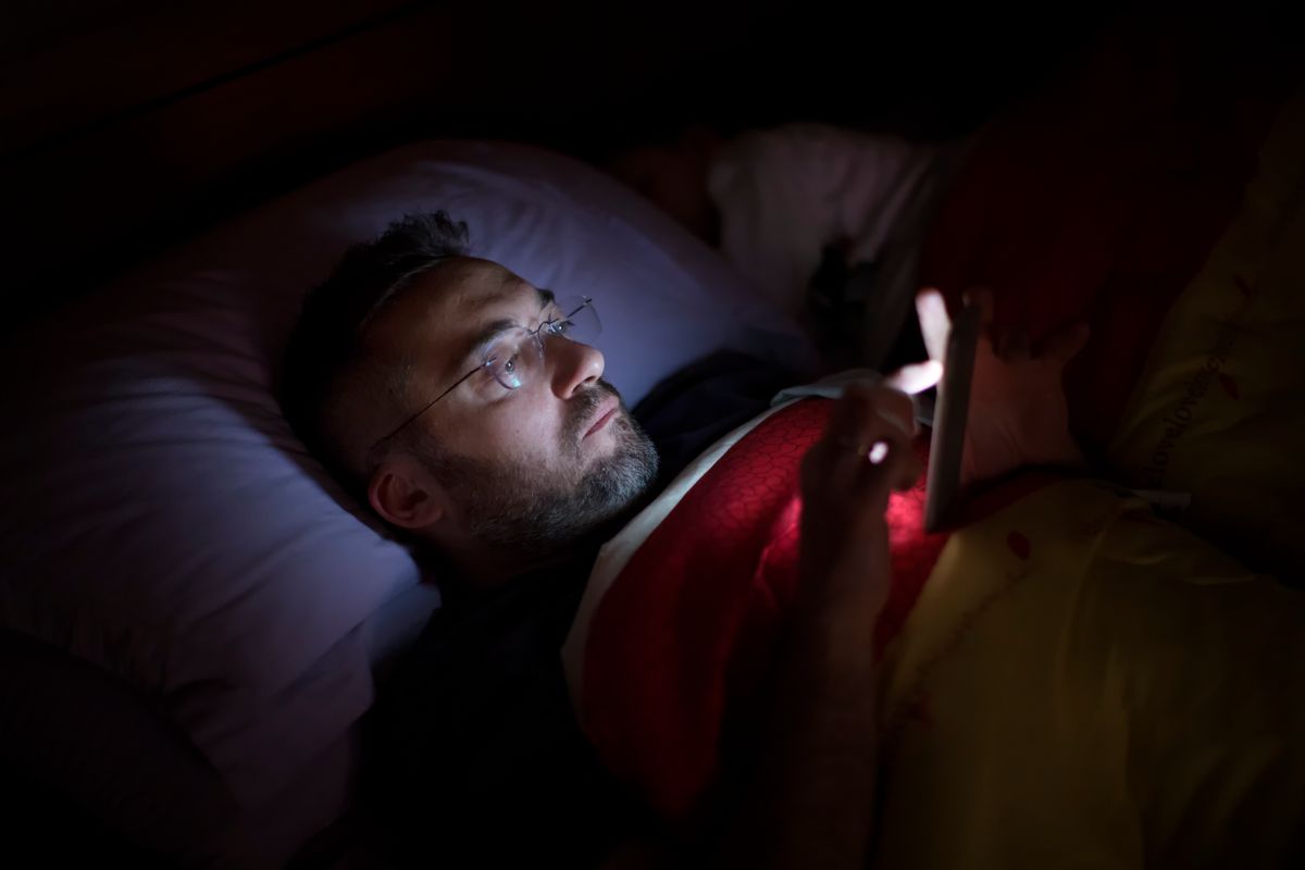A man in glasses lies in bed, illuminated by the soft glow of his smartphone screen. He appears focused and relaxed while scrolling before sleep, wrapped in a blanket.
