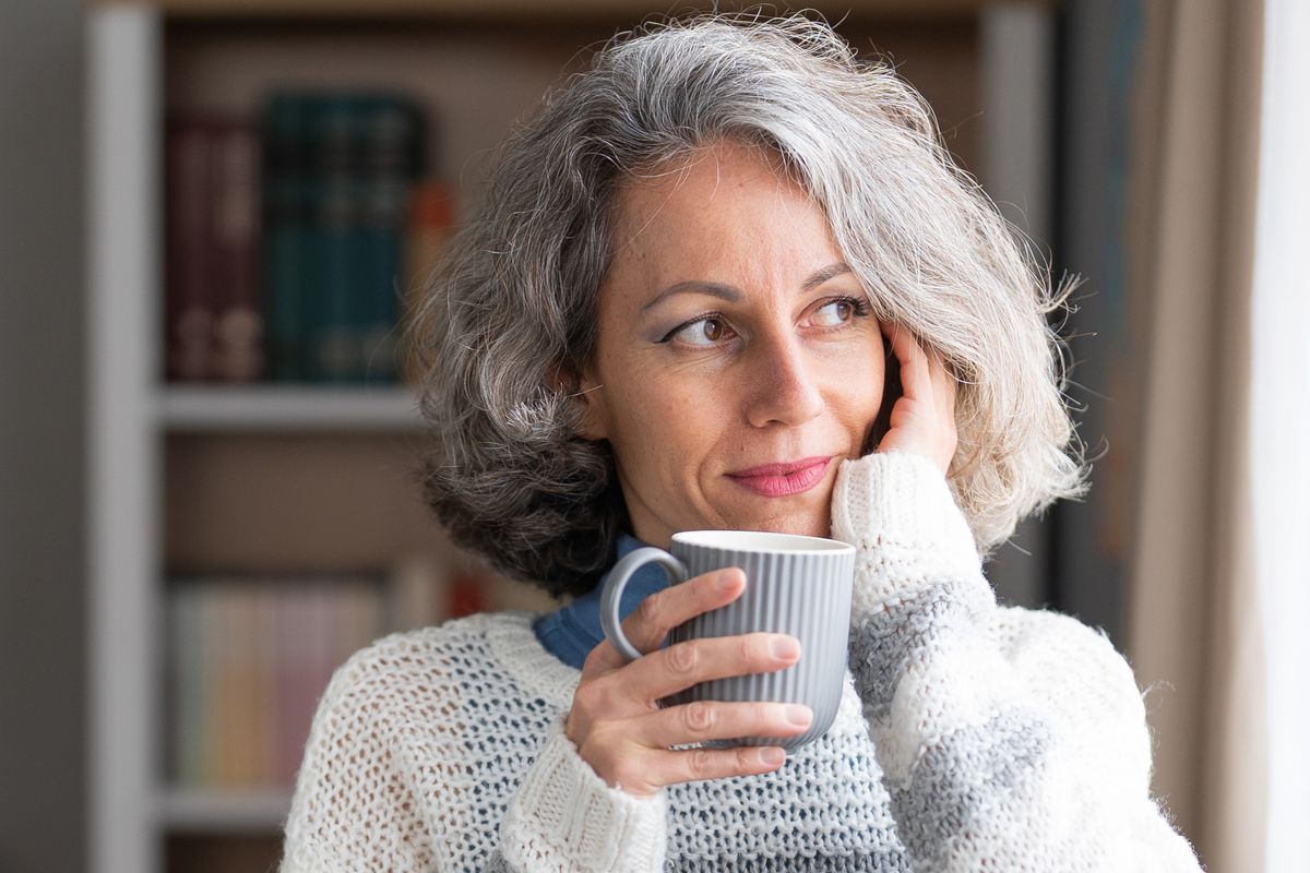 Mature adult woman standing holding a coffee mug, looking away and thinking at home after having iHerb collagen
