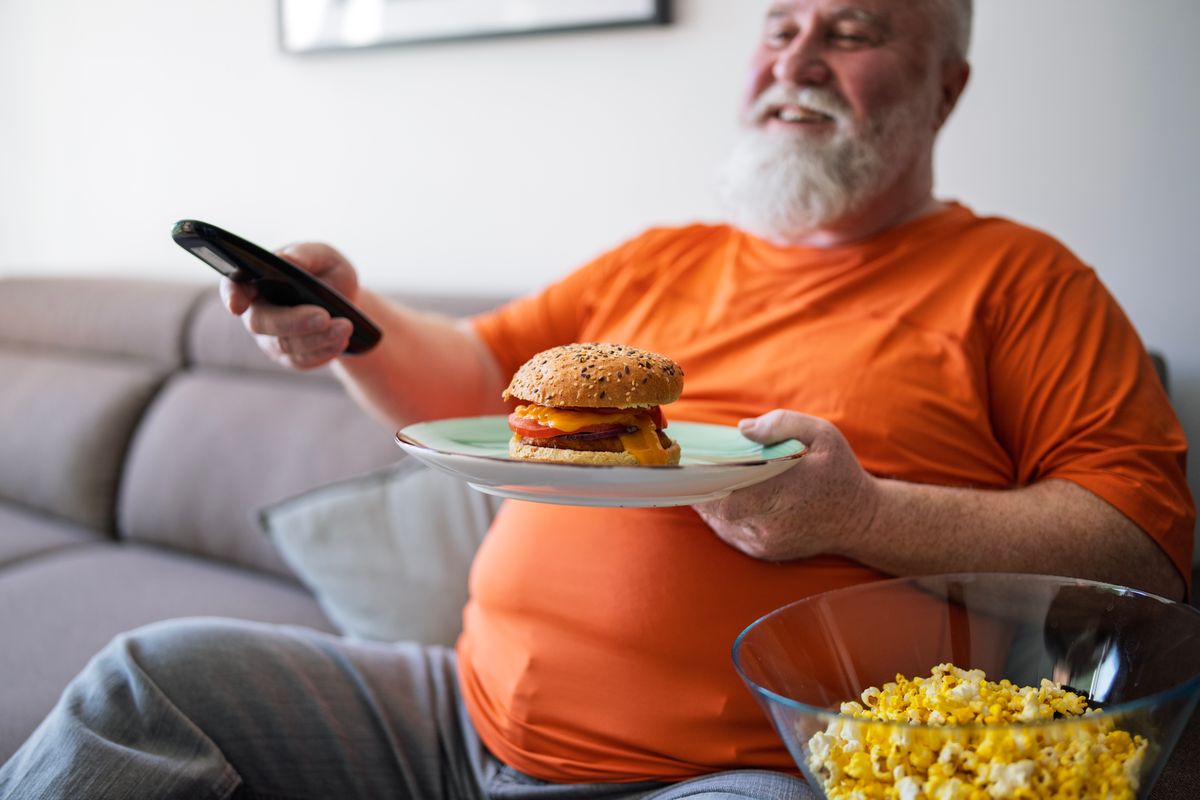 Overweight senior man eating cheeseburger and popcorn on the sofa in front of the TV.