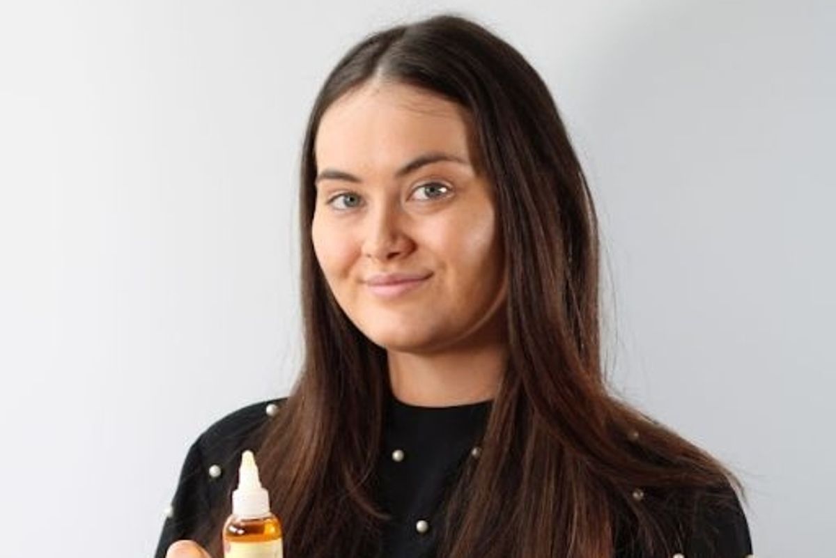 The image shows the young woman with fair skin and brown long hair posing with a bottle of her product and smiling with a blank background in this portrait picture