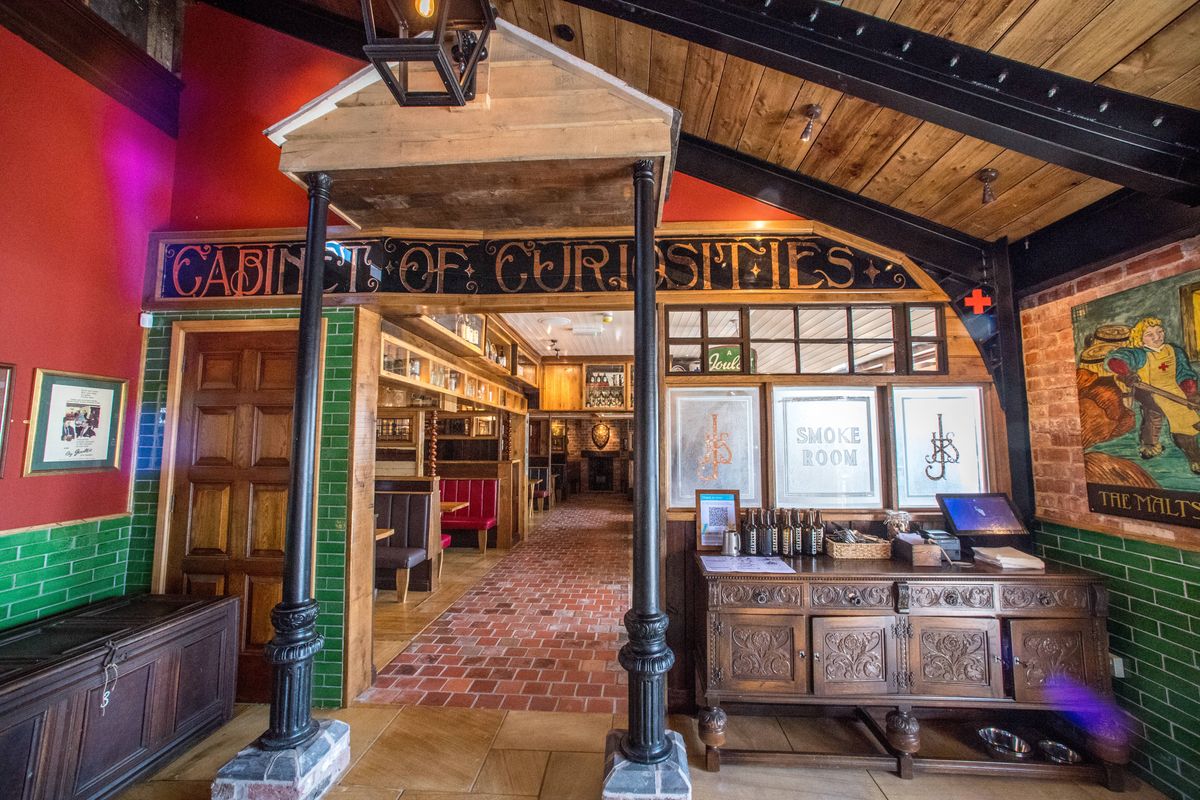 Interior view of a decorated room with wooden beams, vibrant wall colors, and intricate architectural details. The space features a wooden pillar supporting a canopy, an ornate wooden cabinet with various items on top, and framed artworks on the wall.