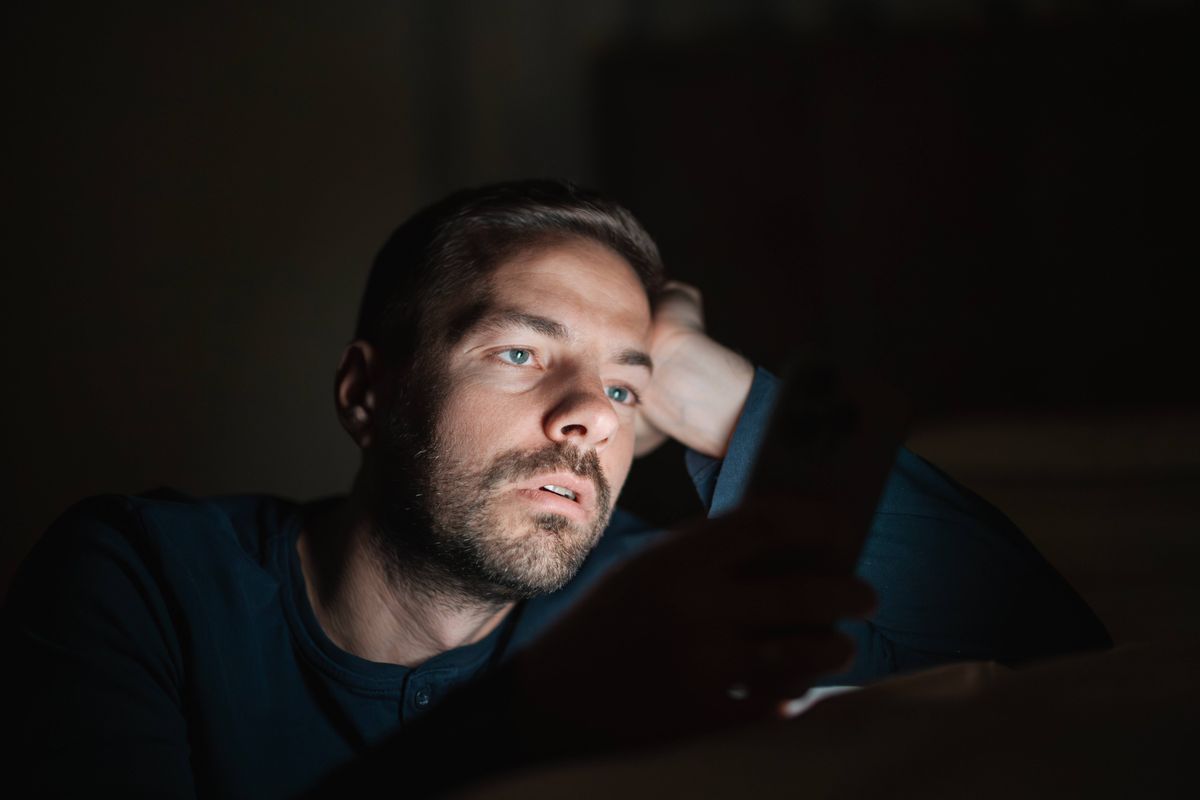 Man with stubble and blue eyes looking intently at a glowing smartphone screen, creating a strong contrast in the dark environment of a bedroom, feeling tired or experiencing insomnia