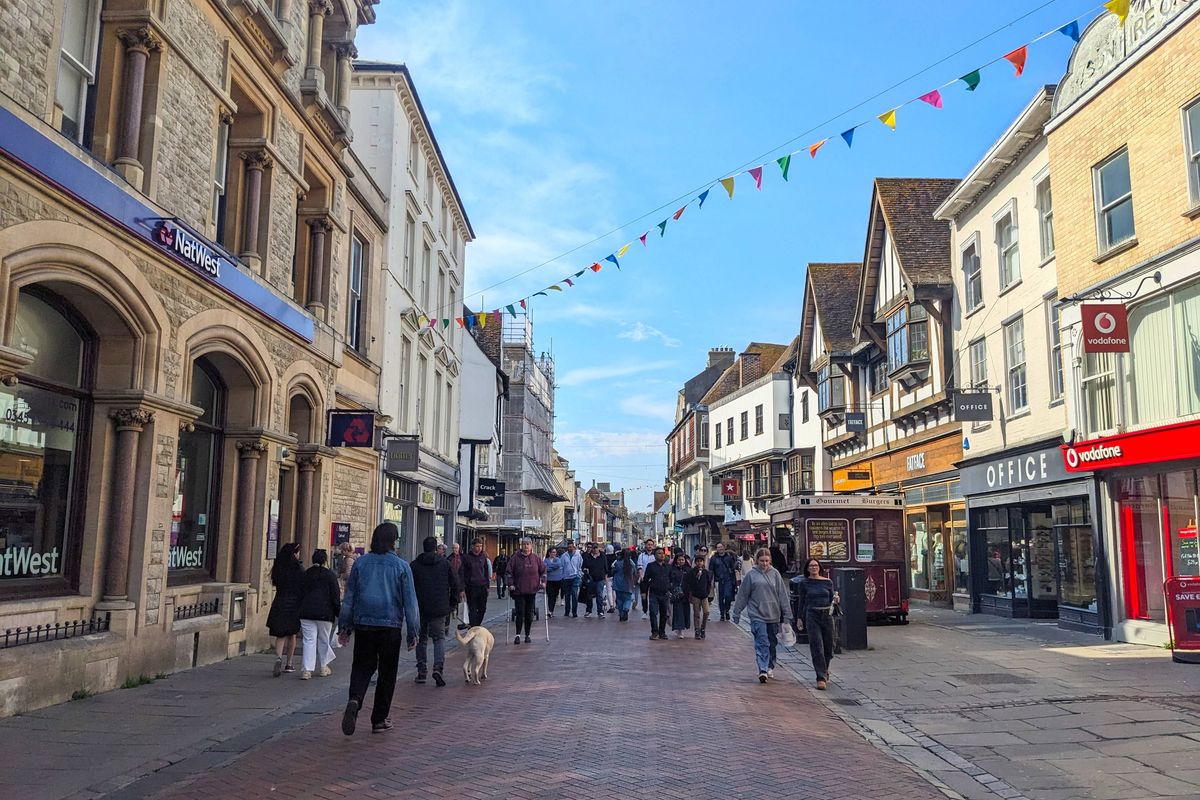 Canterbury, UK - April 19 2025: People walk past traditional architecture and vibrant shopfronts on the bustling Parade adorned with colourful bunting under a sunny blue sky