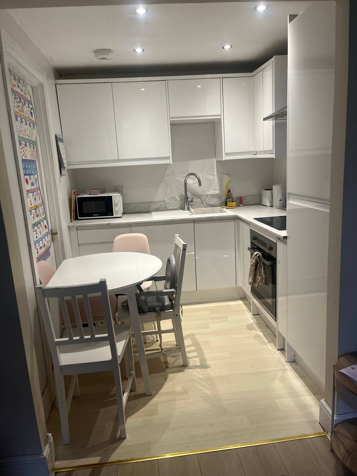 Interior of a contemporary kitchen featuring white cabinetry, a central dining table, and a compact dining set. The kitchen includes a microwave on a countertop, a sink with a faucet, and a built-in oven. The room is well-lit with overhead lighting, and the flooring is made of light-colored tiles.