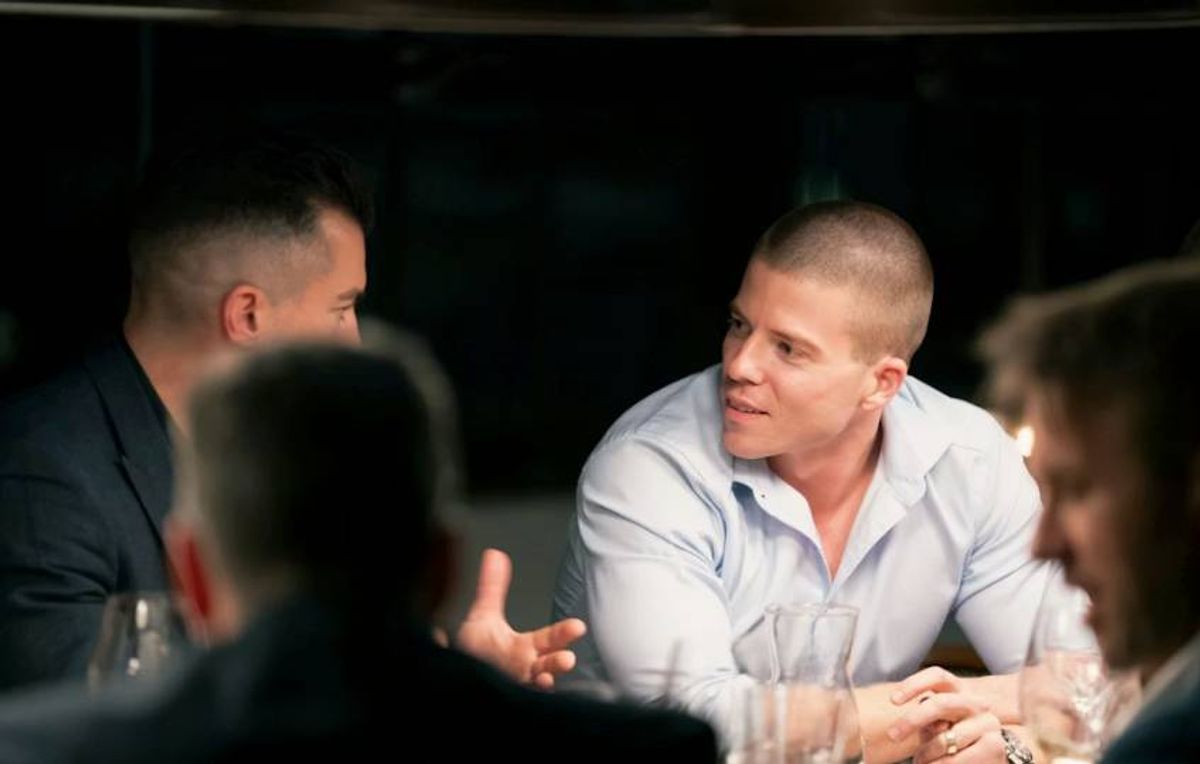 A group of individuals engaged in a discussion, with one individual dressed in a light-colored long-sleeve shirt seated in the center.