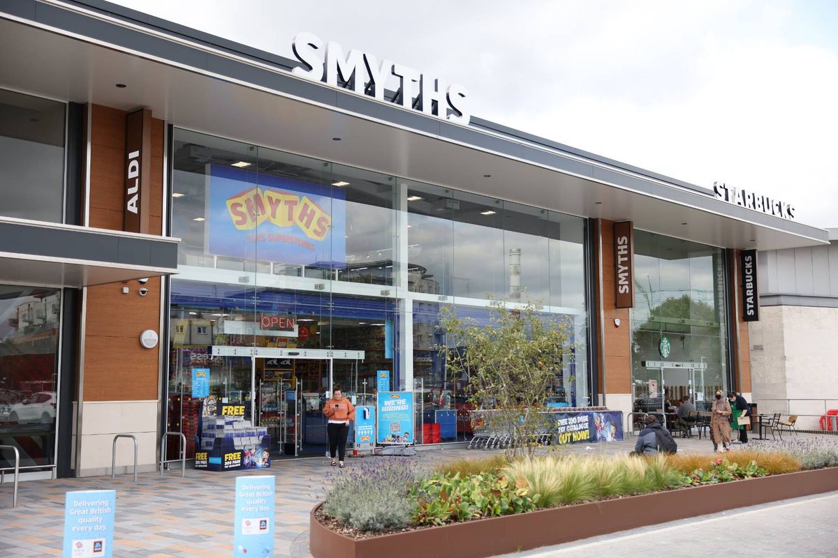 An exterior view of a modern retail establishment features a prominent sign reading "Smyths" with a large glass front showcasing the interior. The entrance is well-lit, and there is a decorative banner hanging above the entrance. Several customers are visible inside, navigating the store's layout. The establishment is surrounded by greenery, contributing to a welcoming atmosphere.