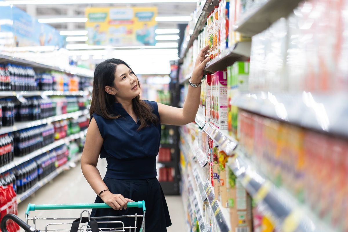 Woman reaching for a product on a supermarket shelf while pushing a shopping cart, browsing beverages in a grocery store.