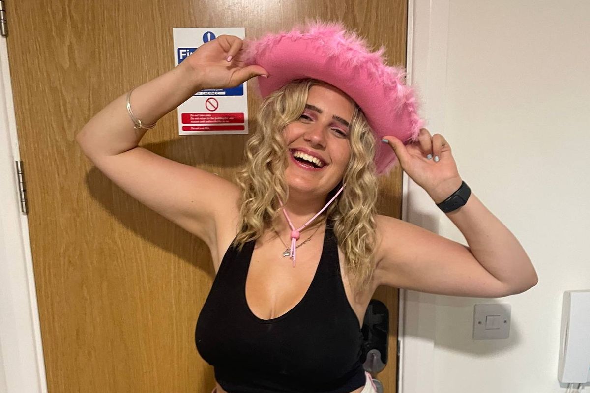 An individual is standing indoors, wearing a black crop top and a pink cowboy hat, with a joyful expression and hands positioned behind the head. The setting appears to be a room with a wooden door in the background.