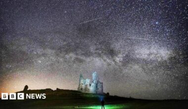 Grey castle ruins are illuminated by thousands of stars in the sky. The milky way is visible, as are shooting stars. In the foreground, there is a silhouette of a man looking up at the sky with a torch.