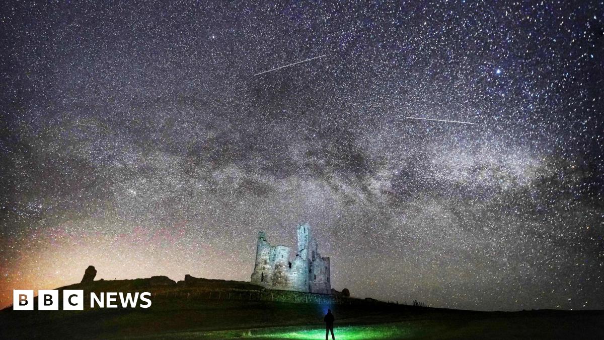 Grey castle ruins are illuminated by thousands of stars in the sky. The milky way is visible, as are shooting stars. In the foreground, there is a silhouette of a man looking up at the sky with a torch.
