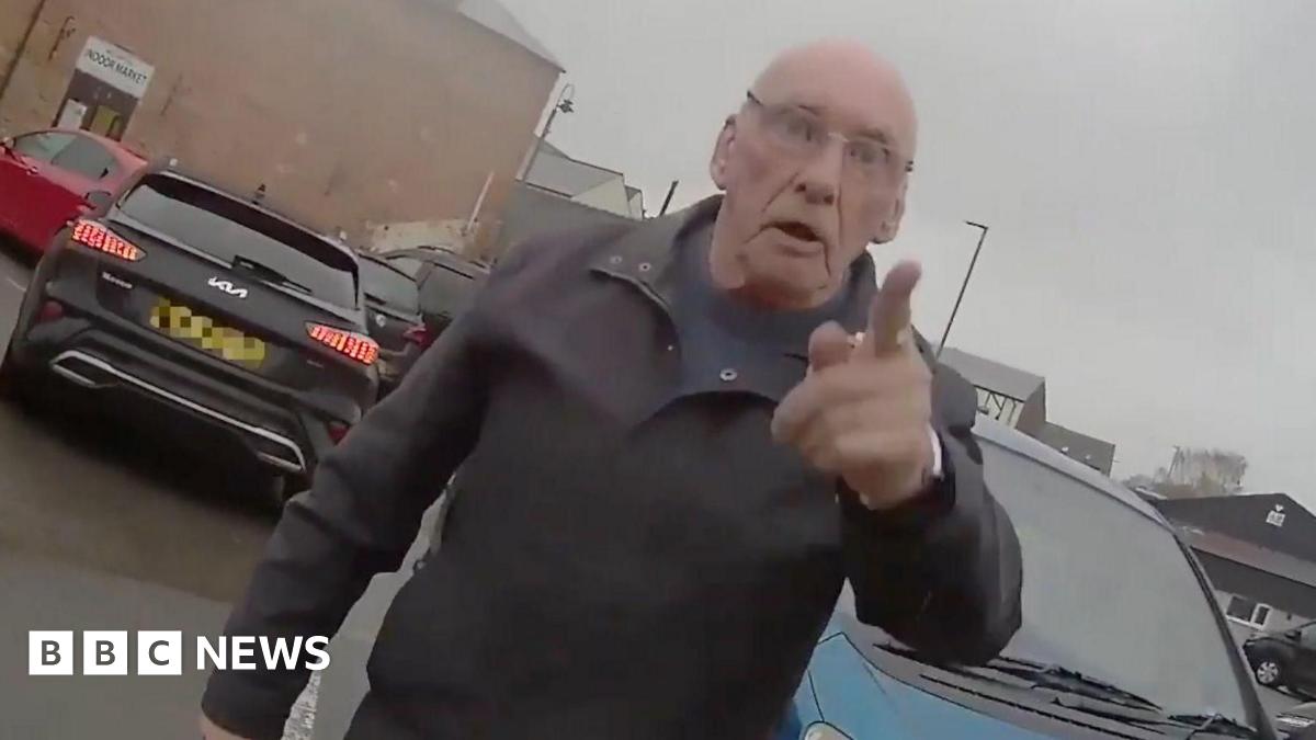 A man standing in a car park. He has his finger pointed at the person wearing the body-cam.