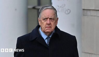William Lloyd-Lavery walking outside. He is wearing a navy jacket over a light blue shirt with a navy tie with multi-coloured spots on it. He has grey hair.