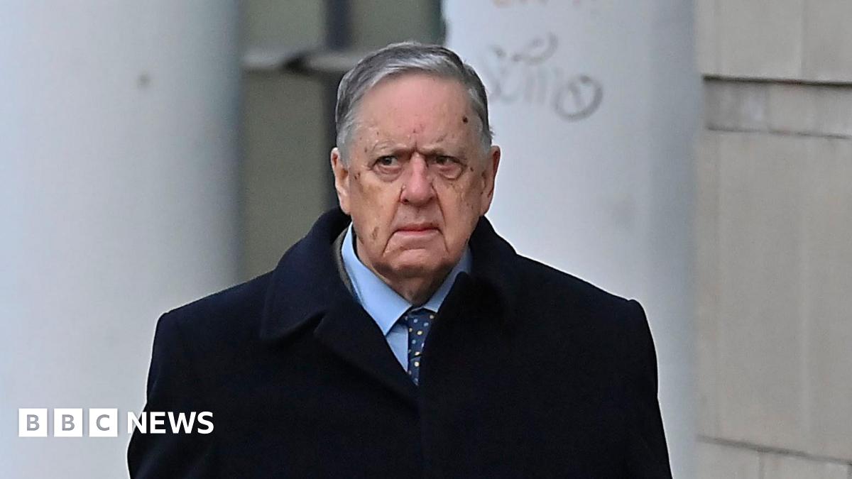 William Lloyd-Lavery walking outside. He is wearing a navy jacket over a light blue shirt with a navy tie with multi-coloured spots on it. He has grey hair.