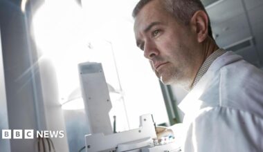 Dr Stephen Grimes, Associate Professor in Stable Isotope Geochemistry and Climate Change at the University of Plymouth. He is stood up and looking to the left. He is wearing a white lab coat. In front of him is a microscope and bright light.