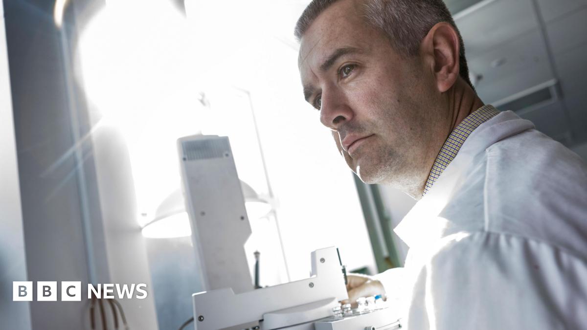 Dr Stephen Grimes, Associate Professor in Stable Isotope Geochemistry and Climate Change at the University of Plymouth. He is stood up and looking to the left. He is wearing a white lab coat. In front of him is a microscope and bright light.