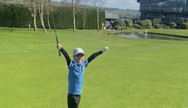 Freddie wearing a light grey cap and blue gold top and black golf pants and white golf shoes throws his arms up in the air to celebrate his hole-in-one on Carus Green Golf Club course.