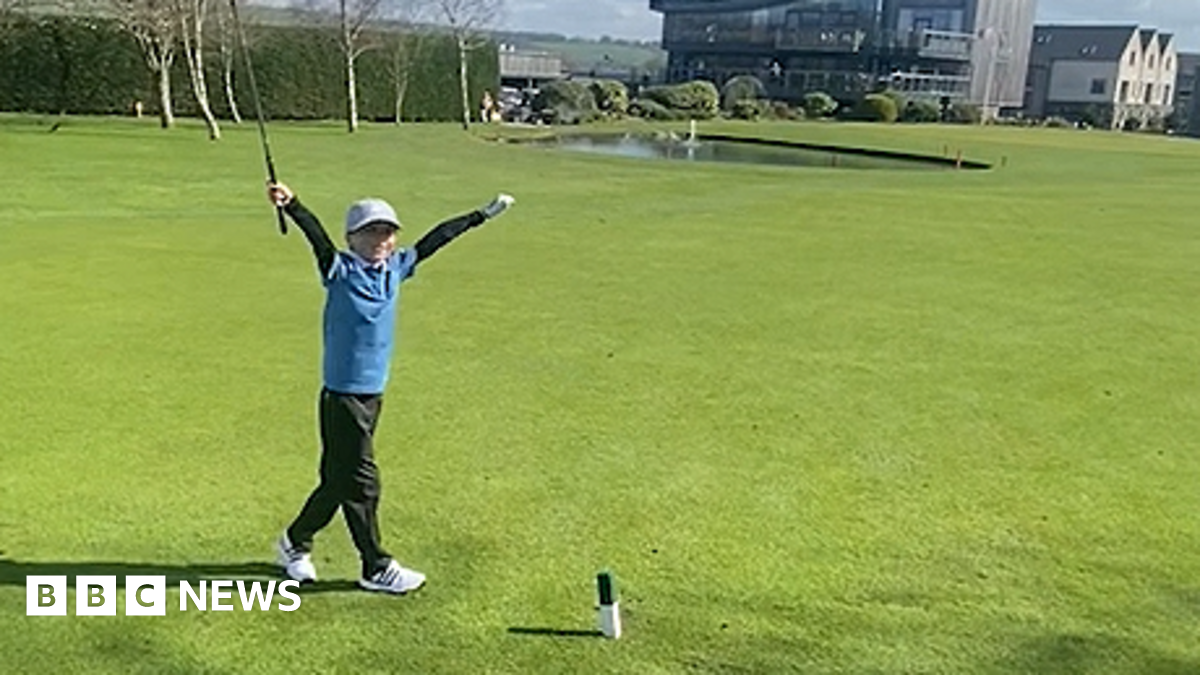 Freddie wearing a light grey cap and blue gold top and black golf pants and white golf shoes throws his arms up in the air to celebrate his hole-in-one on Carus Green Golf Club course.