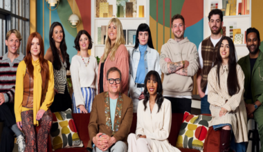 Alan Carr is sat down on a red settee wearing a brown jacket and grey trousers. He is sat next to judge Michelle Ogundehin, who is dressed in a cream jacket and matching skirt. They are joined by ten people, who are all contestants on the series Interior Design Masters. Sophie from Stoke on Trent is among the contestants and is second left in the photograph. She has long red hair and is wearing a yellow shirt, patterned red trousers and a brown gilet. Ben, from Kinver, is stood behind the red sofa with his arms folded, wearing a beige hoodie.