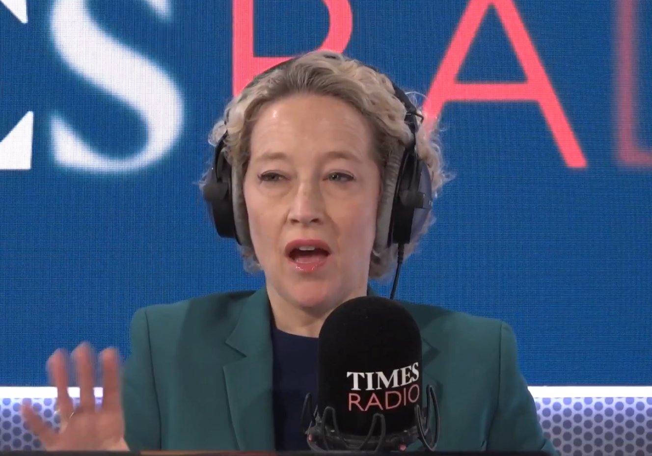 A woman with short curly blonde hair and headphones speaks into a microphone for Times Radio.