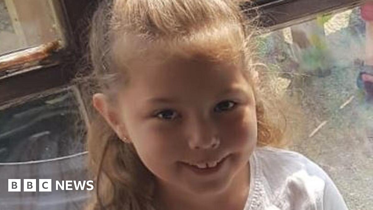 The late Olivia Pratt-Korbel smiles for the camera. She is sat down, back leaning against a window. She has her hair tied up and wears a white shirt.
