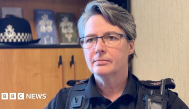 Louise Kennaugh has short dark and grey hair and wears glasses. She's in black police uniform sitting in an office, with a wooden cabinet behind to the left with her police hat resting on a shelf.