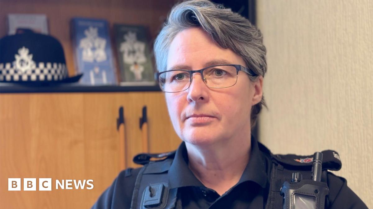 Louise Kennaugh has short dark and grey hair and wears glasses. She's in black police uniform sitting in an office, with a wooden cabinet behind to the left with her police hat resting on a shelf.