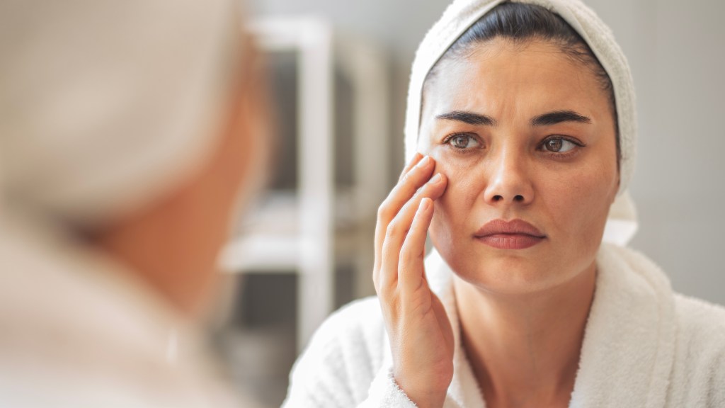 A woman in a white robe and hair towel touches the skin around her eye, looking closely at her reflection in the mirror with a worried expression.