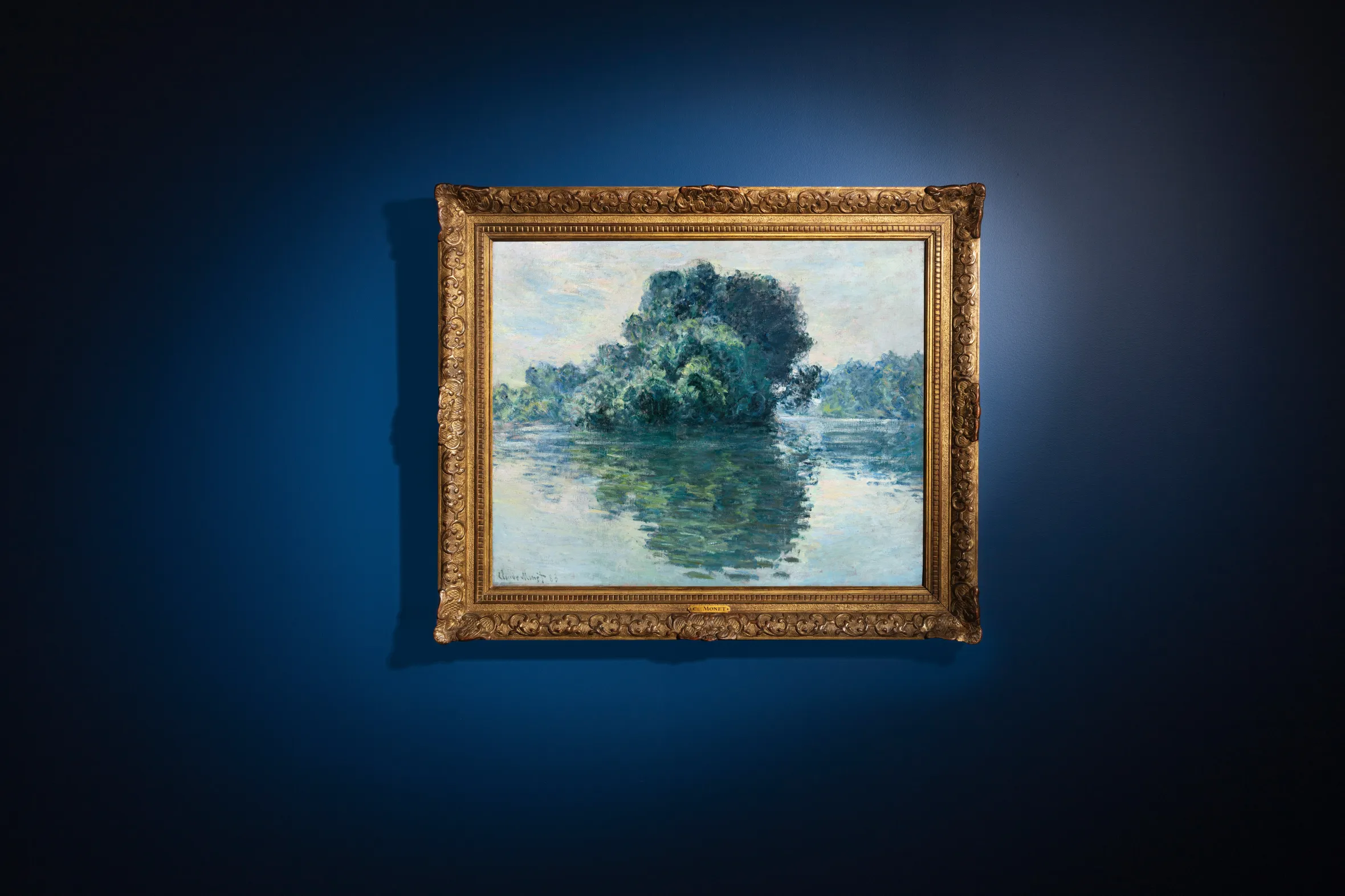 Claude Monet's painting "Les îles de Port-Villez," a landscape of trees reflected in water, in an ornate gold frame on a dark blue wall.