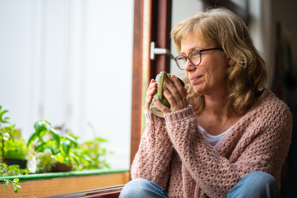 An older woman with blonde hair and glasses sips tea, looking pensively out a window.