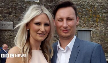 A couple pose for a picture - the woman on the left has long blonde hair and a light-pink dress. The man has short brown hair, a blue jacket and white shirt. There is an old stone building behind them.
