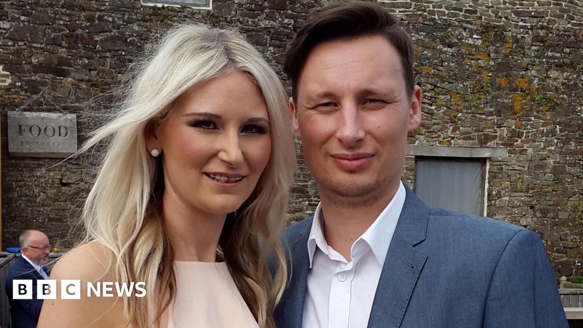 A couple pose for a picture - the woman on the left has long blonde hair and a light-pink dress. The man has short brown hair, a blue jacket and white shirt. There is an old stone building behind them.