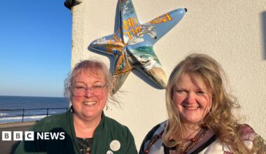 A woman with a blonde fringe and hair tied back is wearing large clear-rimmed glasses and a green cardigan and is smiling into the camera. Next to her is a woman with long blonde hair also smiling into the camera. Behind them is a wall with a starfish sculpture which says 'Lake-Land By The Sea' in yellow lettering with a background of the seaside with seagulls, sky, sand and the sea. The actual sea can be seen behind them.