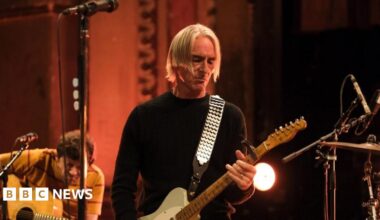 Paul Weller playing a guitar, with blond hair in a centre parting, looking down at the guitar. In the background is another guitarist and the edge of a drumkit.